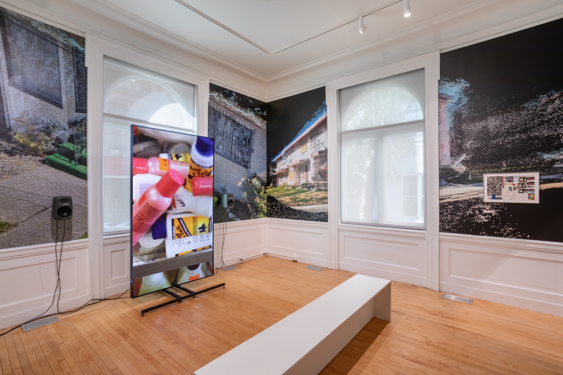 Installation view of large monitor with film still of bottles to the left and framed collage work on wall to right with film-still wall paper behind