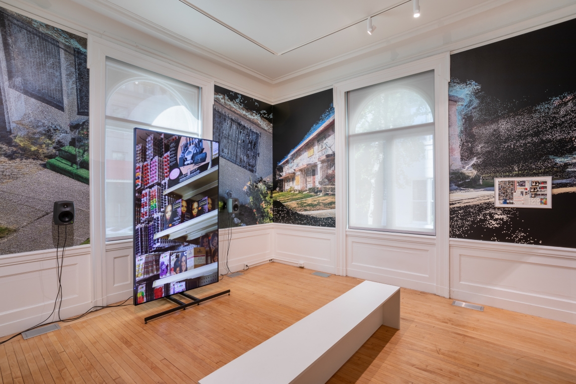 Installation view of large monitor to the left with film still of store shelves and a framed collage work on wall to right with film still wallpaper behind