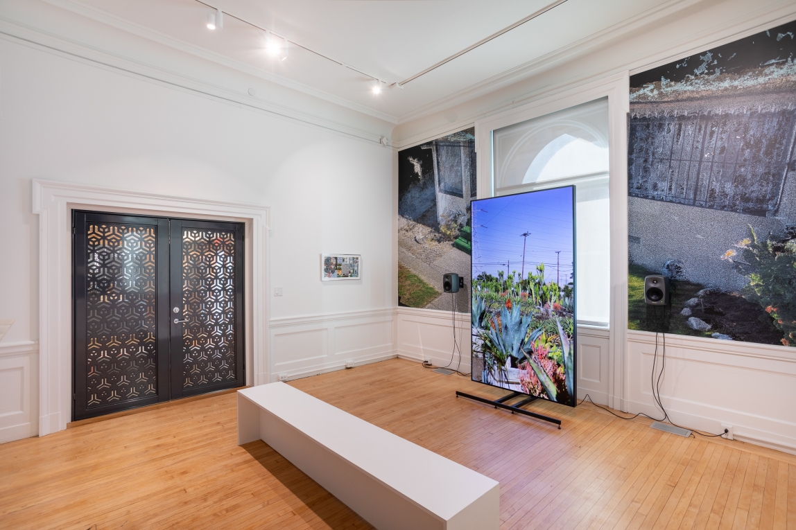 Installation view of gallery with large monitor to the side with film still of landscape and framed collage on far wall next to closed metal doors