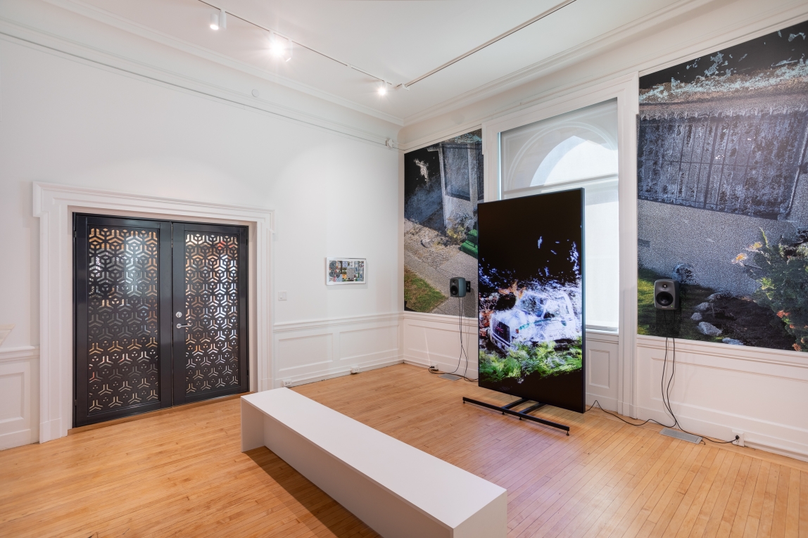 Installation view of large monitor with graphic landscape on screen, framed collage work on far wall next to closed metal doors