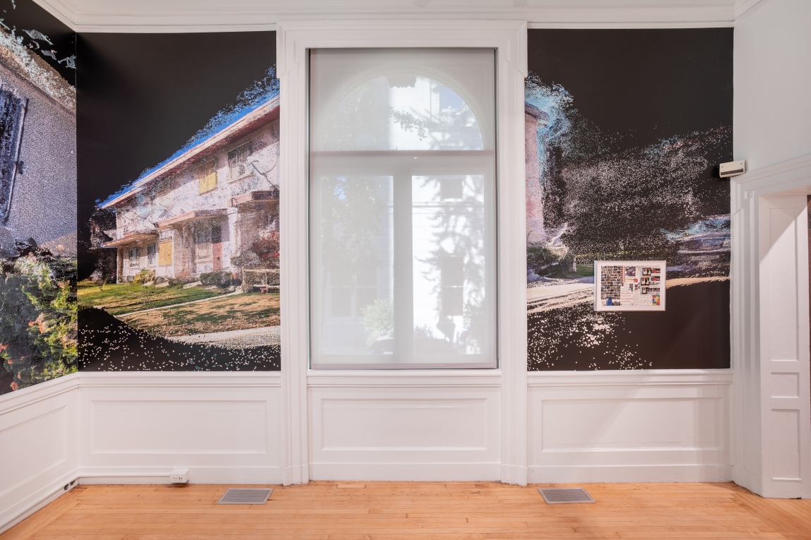 Installation image of wall paper surrounding a window featuring a film still of a building with a framed collage work on top to the right