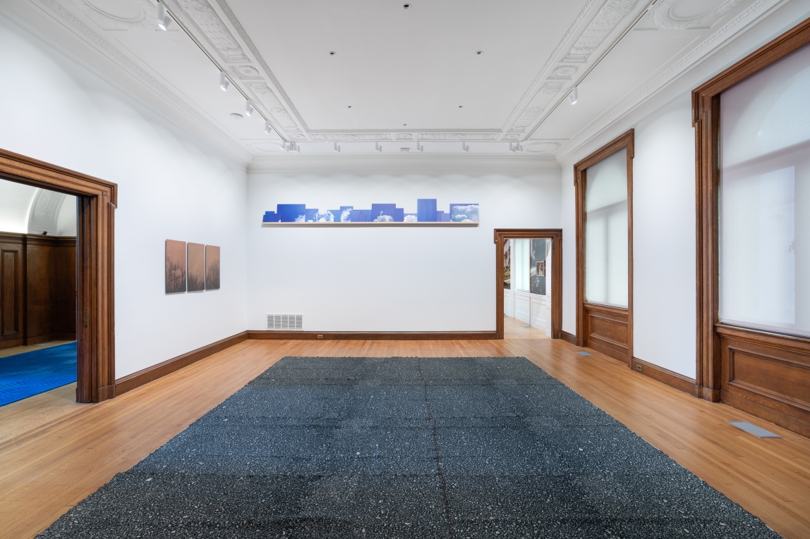 Installation view of gallery with copper printed artwork on one wall, images of blue sky on a shelf on another and a large textile work occupying the center floor space of the room