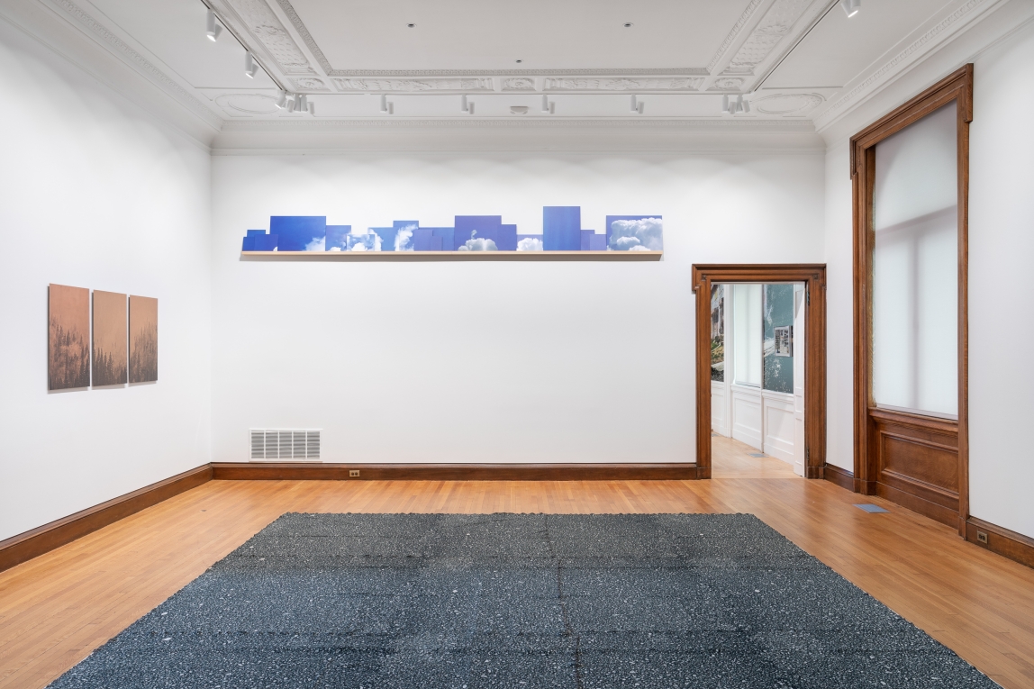 Installation view of gallery with a series of sky images on a shelf on back wall, copper printed landscape plates on right wall and large textile work on floor in foreground