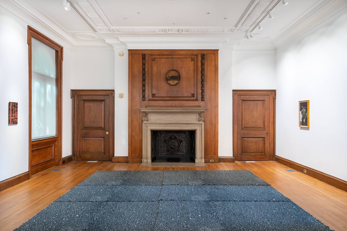 Installation view with large textile work on platform on floor, fireplace in background and a landscape work hanging on the walls facing one another