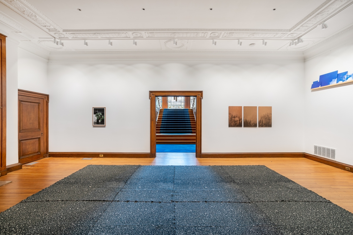 Installation view of gallery with large textile work on floor pedestal in center fo room, and three other works hanging on the back and side wall
