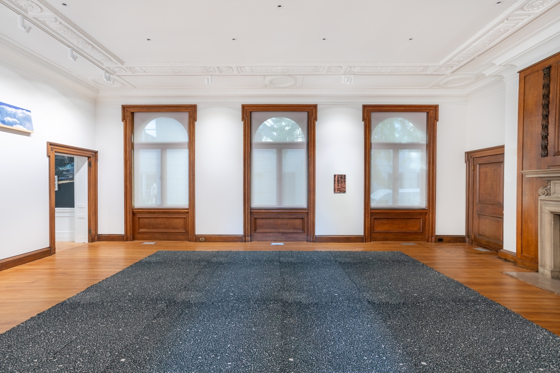 Installation view of gallery with three large windows in background, on artwork hanging in-between two of them, and large textile work in center floor of room