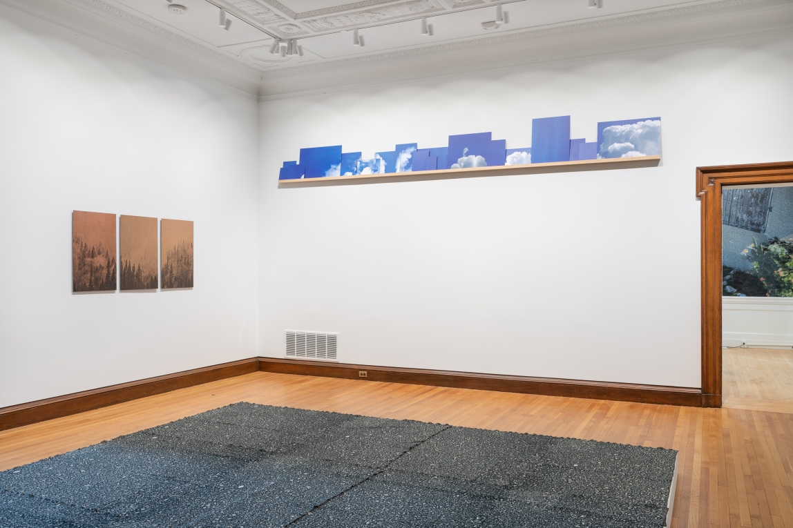 Installation view of corner of gallery with three copper panels hanging on one side, a shelf of sky images hanging no other and textile floor work in foreground
