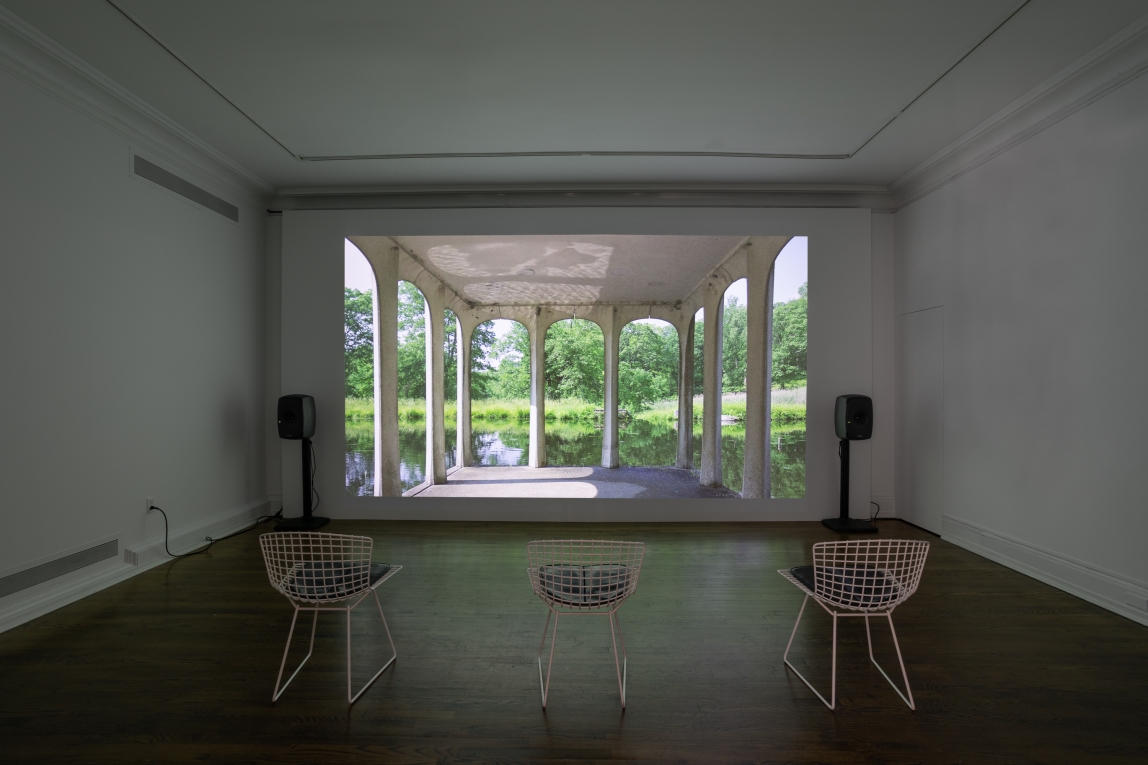Installation view of a film still projected on back wall featuring a portico and landscape with three chairs lined up for viewing
