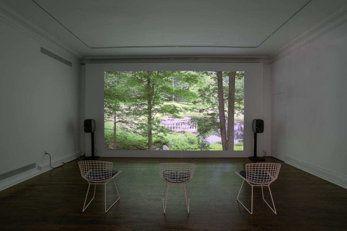 Installation view of film still projected on wall featuring water and trees landscape with three chairs lined up for viewing