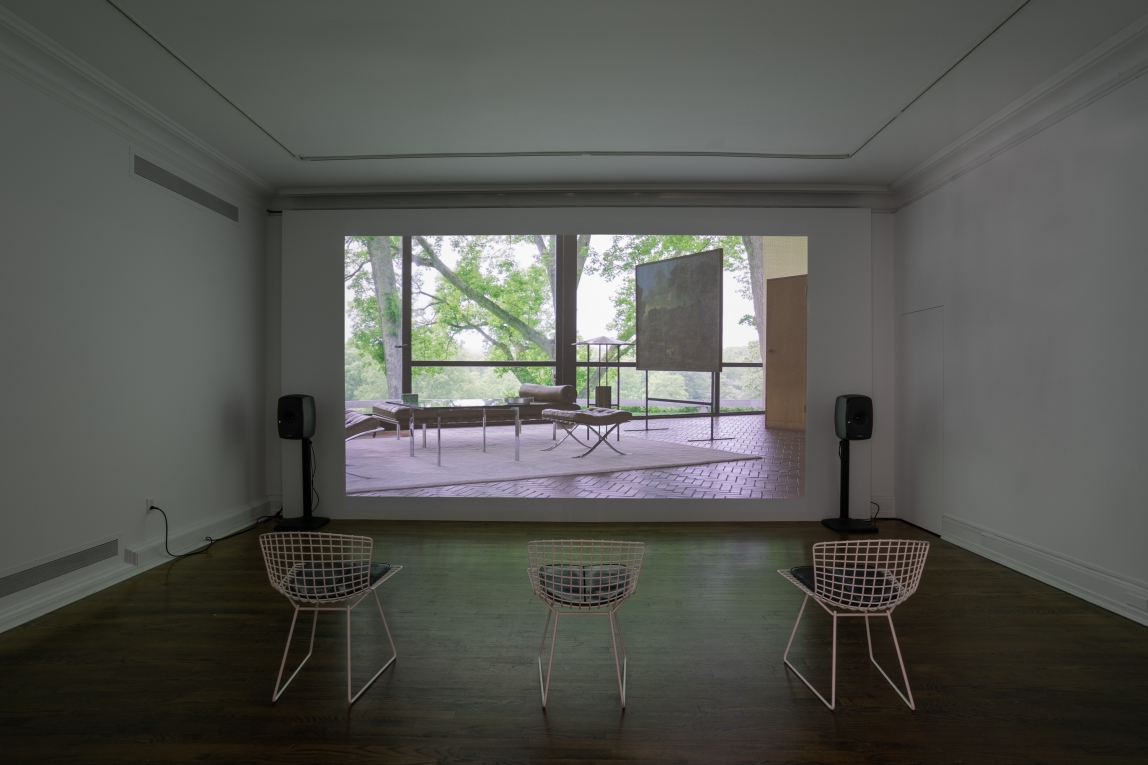 Installation view of film still projected on back wall featuring the Interior of the glass house with three chairs in front for viewing