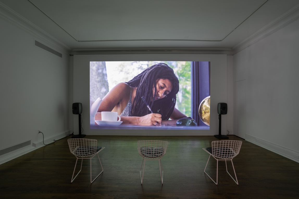 Installation view of film projected on back wall featuring a Black woman writing