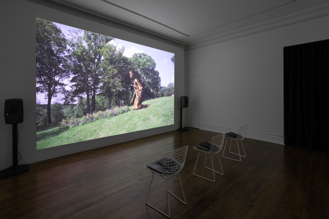 Installation view of film projected on back wall featuring a large Black giant kneeling in a garden