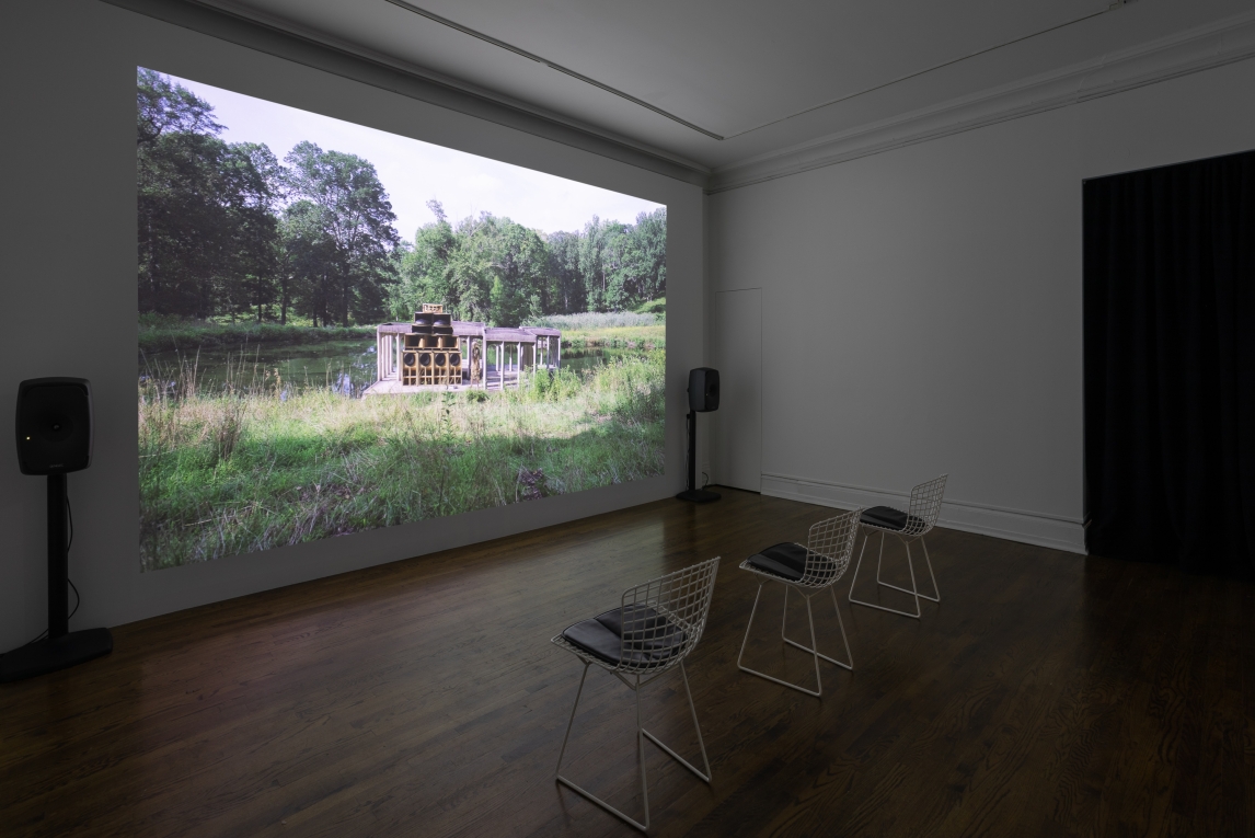 Installation view from angle showing film projected on back wall featuring a portico/building on a watery landscape