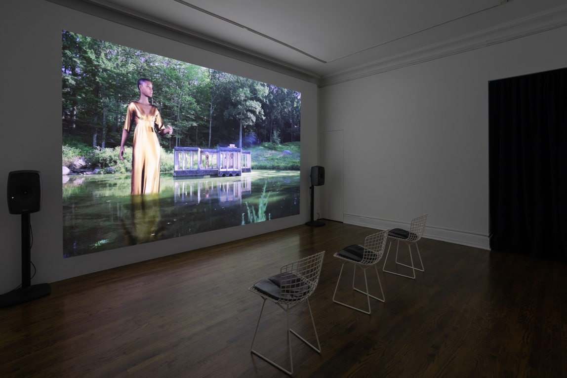 Installation view of film projected on gallery wall, showing a Black giant standing in water towering over a building
