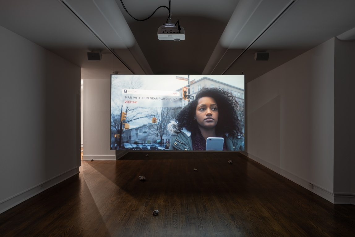 Installation view of film projected in gallery showing a black woman looking out, holding her phone