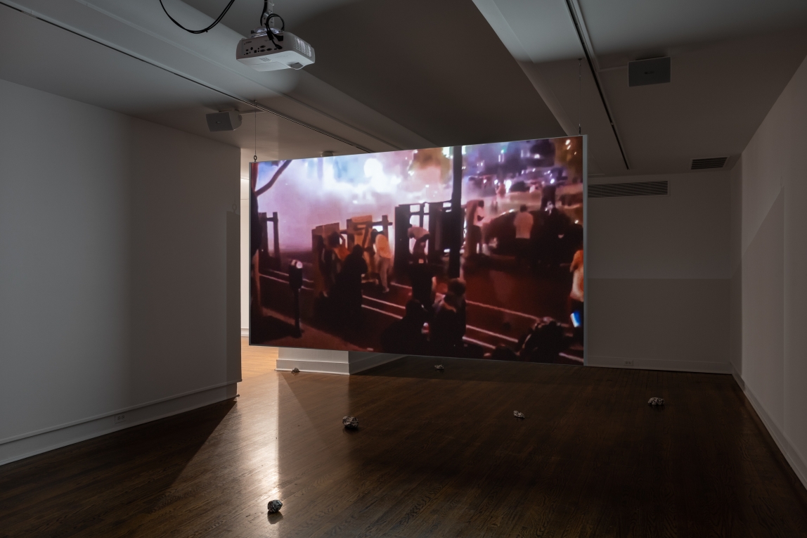 Installation image of film projected in gallery showing a street scene at night with smoke and fire and people behind shields and structures