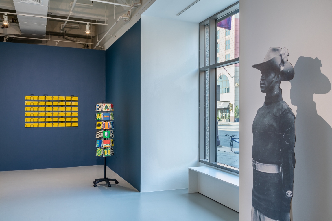 Installation view of painted wall, postcards and cardboard cutout of a Black soldier