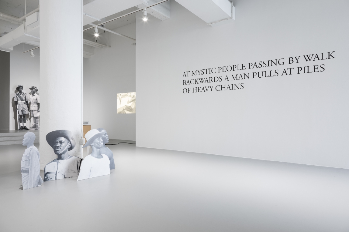 Installation view of text on a wall, projection in the background and cardboard cutouts depicting Black soldiers