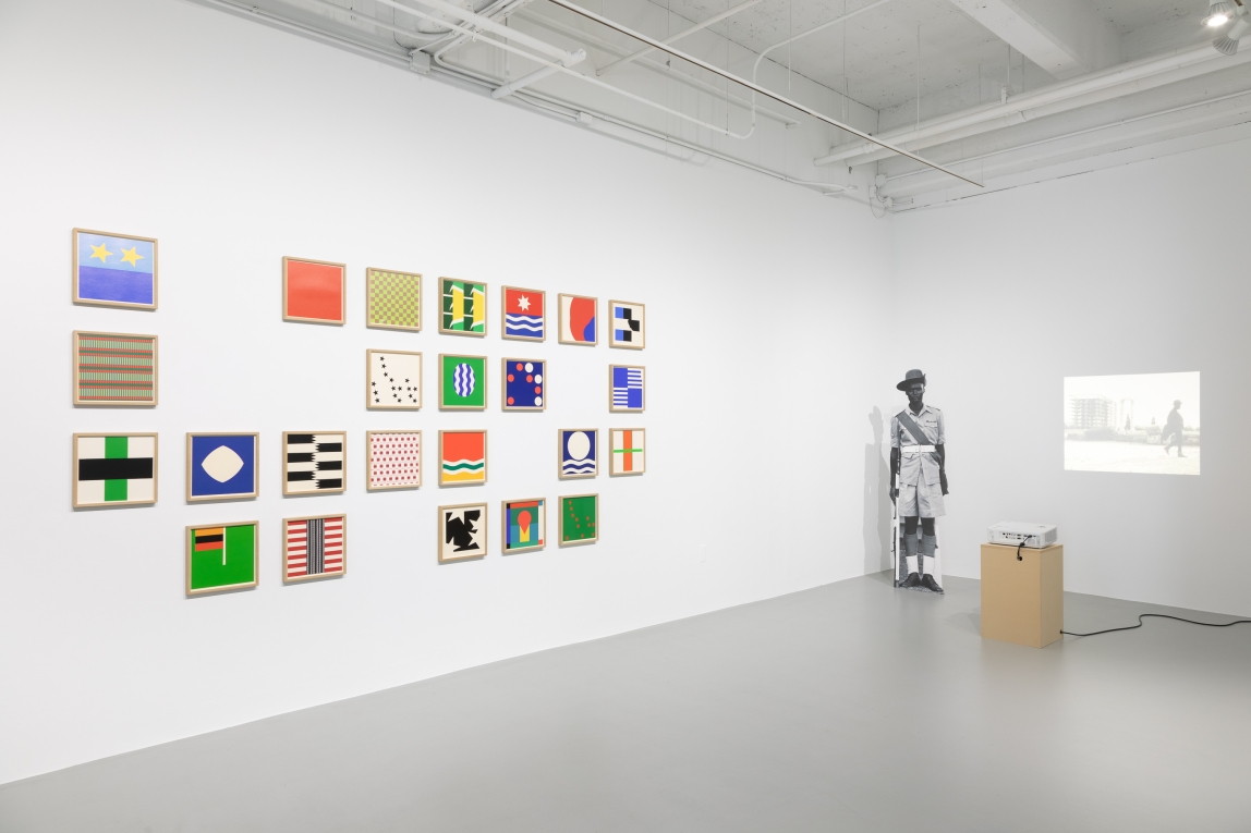 Installation view of bright framed square symbols on left wall, a projection on the right and cardboard cutout of a Black Soldier