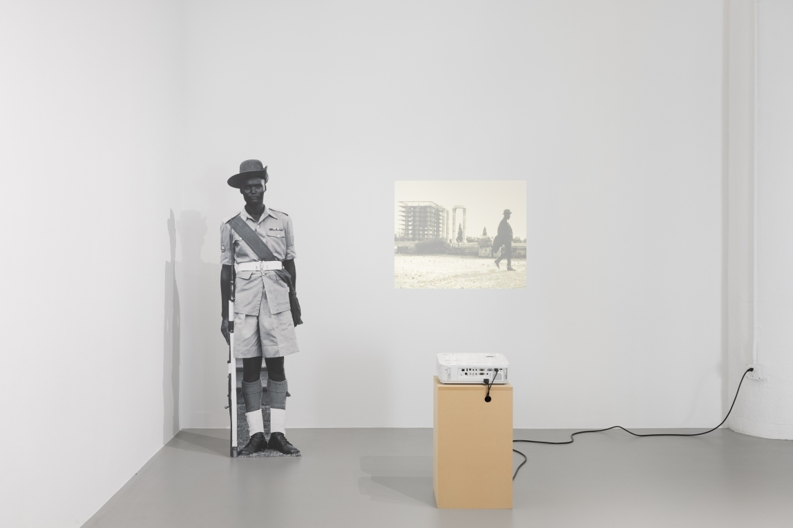 Installation view of a carboard cutout of a Black soldier next to a projection on the wall