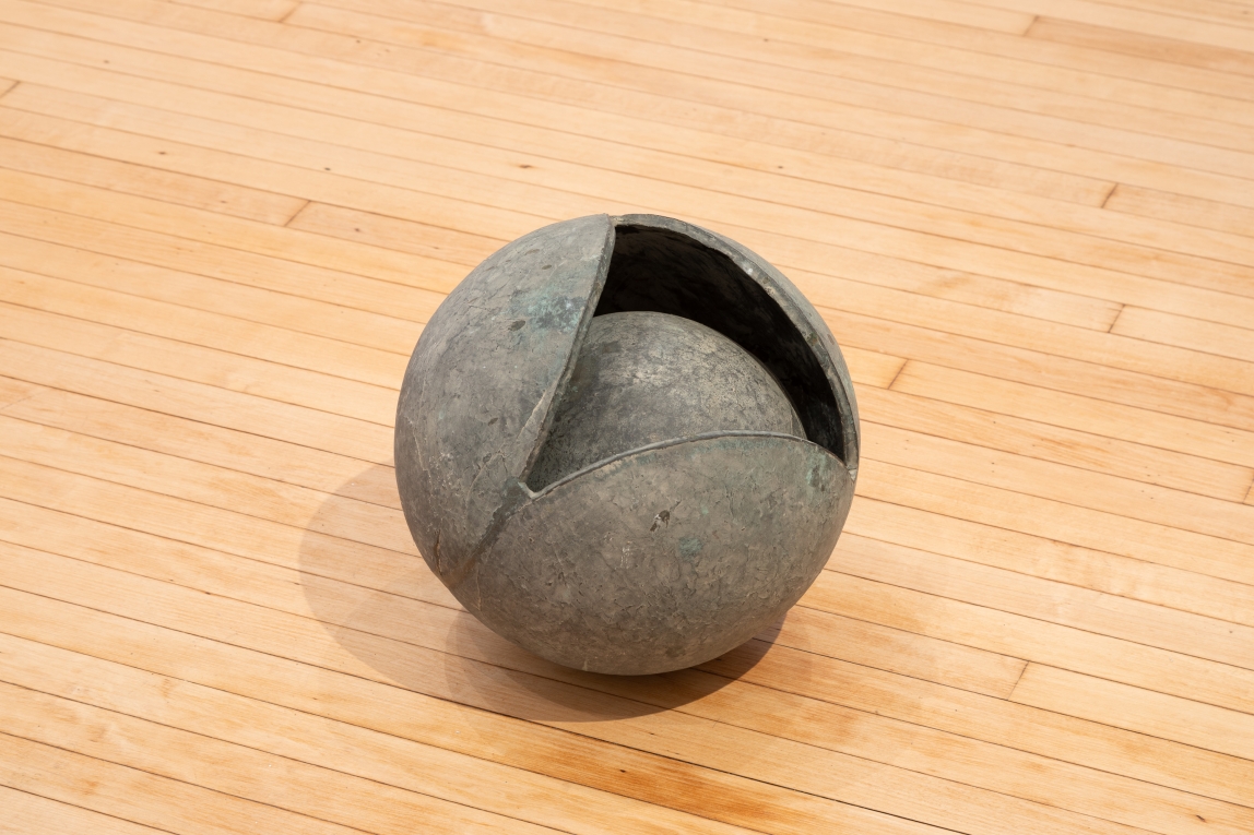 Installation view of metal sculpture on ground showing a sphere within a sphere