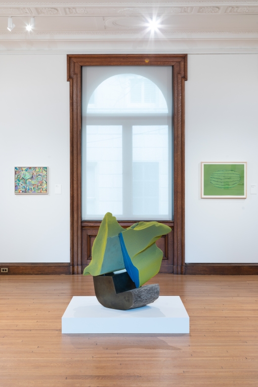 Installation view of two bright paintings on either side of a window and large green and blue sculpture in foreground