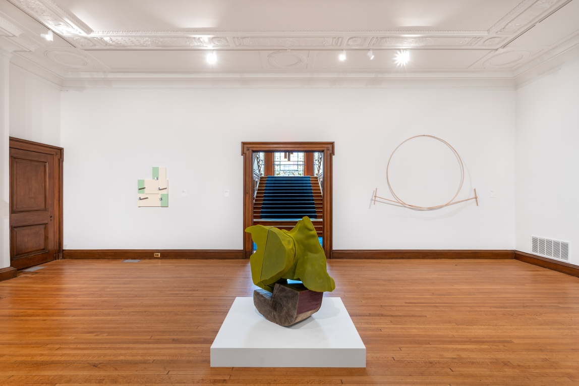 Installation view of large green sculpture in center of gallery and two works hanging on either side of doorway