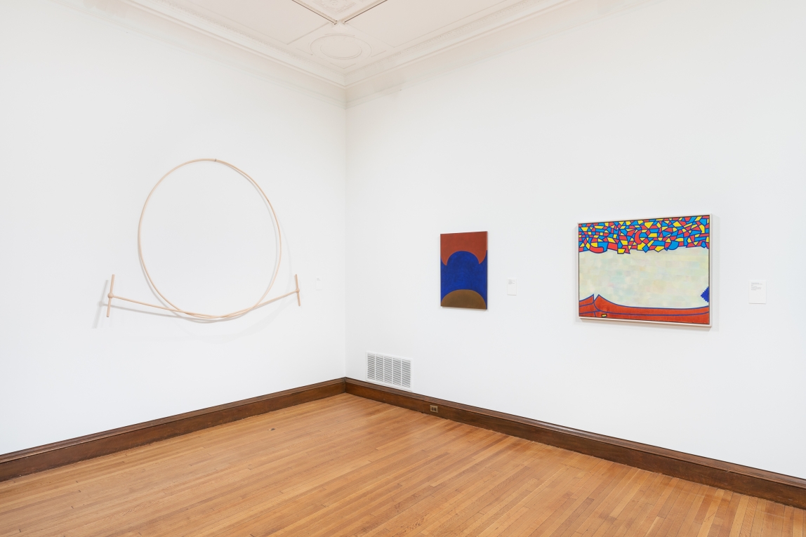 Installation view of two paintings on one wall and a circular wood work on adjoining wall