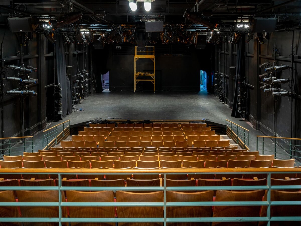 interior of arts bank theater in april 2021. the large theater hall is seen from the rop of the raked seating straight onto the stage area. the walls and stage are black, illuminated by fluorescent lights. spotlights flank the walls. 