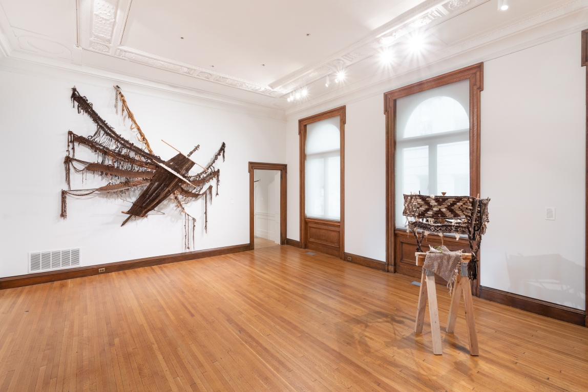 Installation view of two woven fiber artworks in foreground and large windows in background