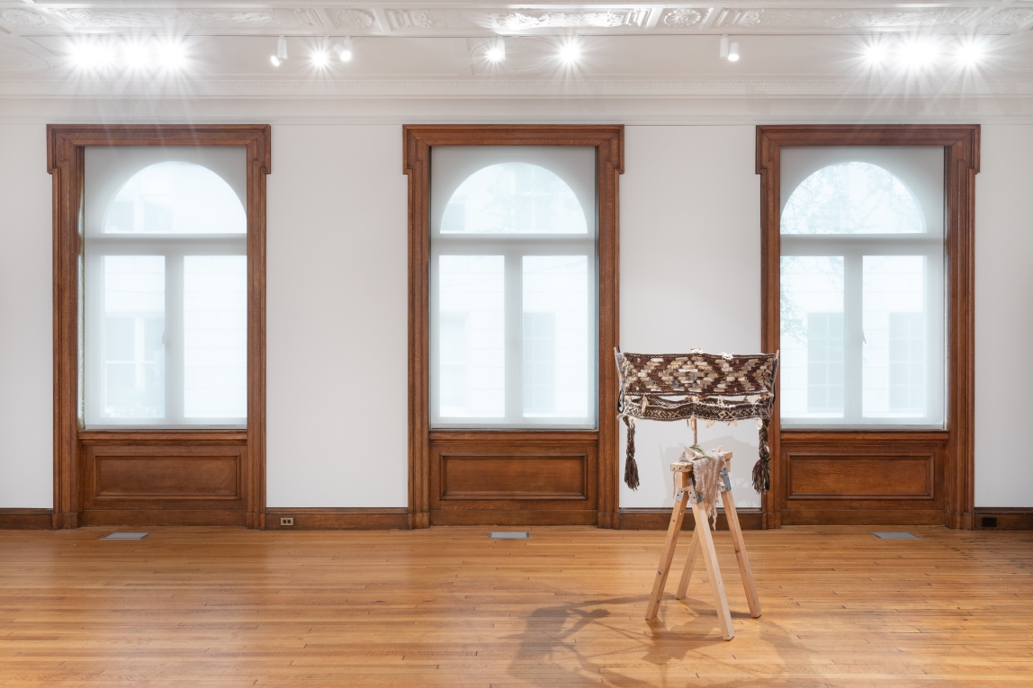 Installation view of woven fiber artwork with wood base in forground, large windows in background