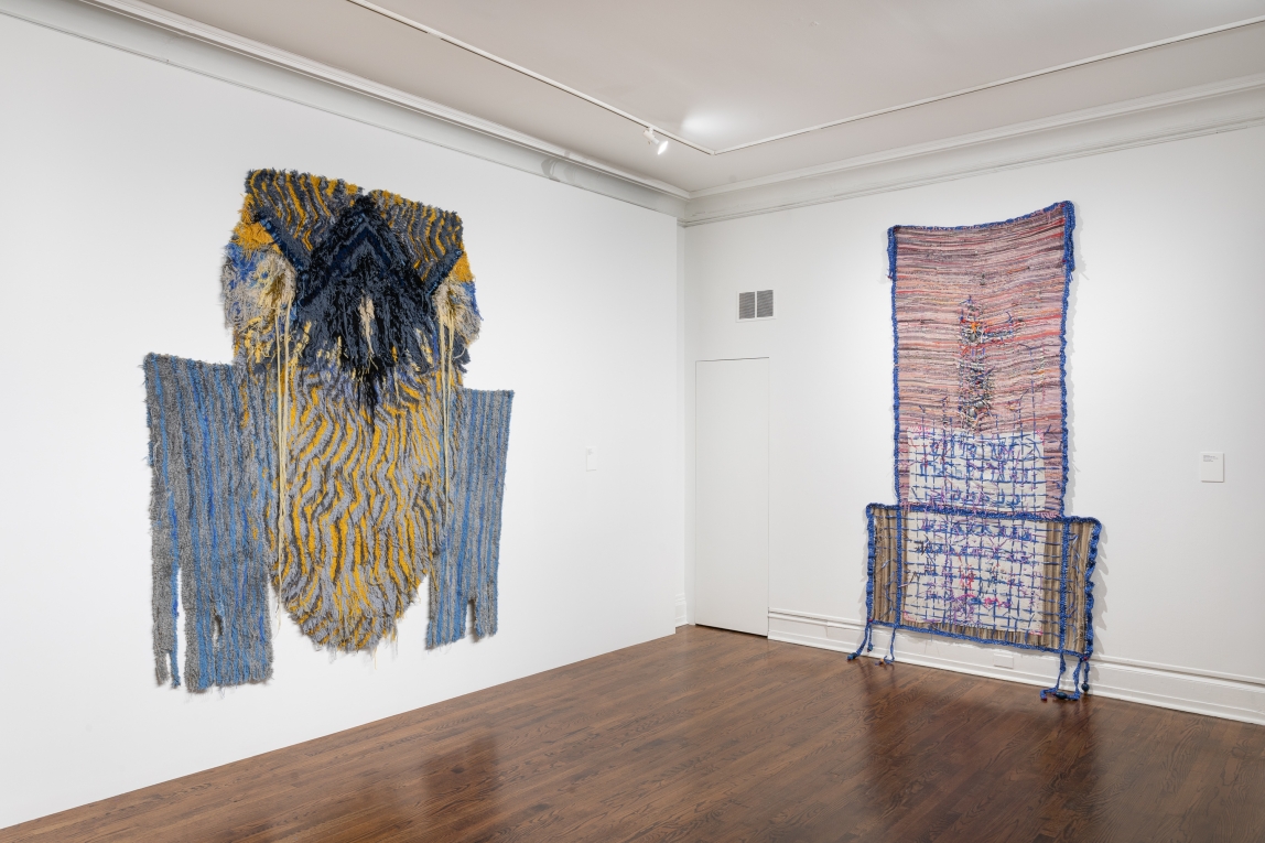Installation view of two large fiber works, a tufted one to the left and a woven mixed fiber work to the right