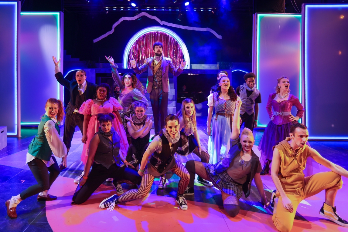fifteen actors pose in semi-crouched stances in a neon and LED-lit stage setting, flanked with neon-ringed panels and accent lights. a violet hue dominates, with a warm glow lighting up the actors' faces.  