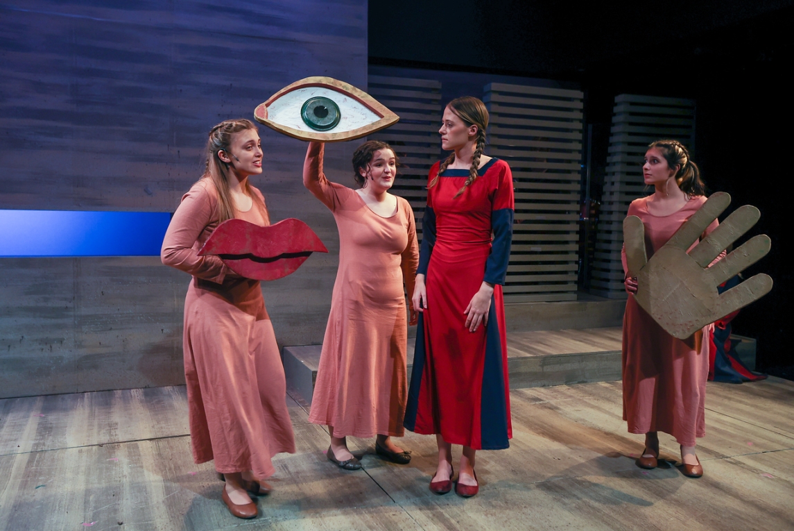 four actors in pink and red dresses stand on a gray wooden stage under bright, even lighting, holding a selection of body parts. a person on the left is holding large red lips at chest level. the next person to the right is holding a large eye aloft in one hand. the person at the far right holds a simple hand with all fingers outstretched. 