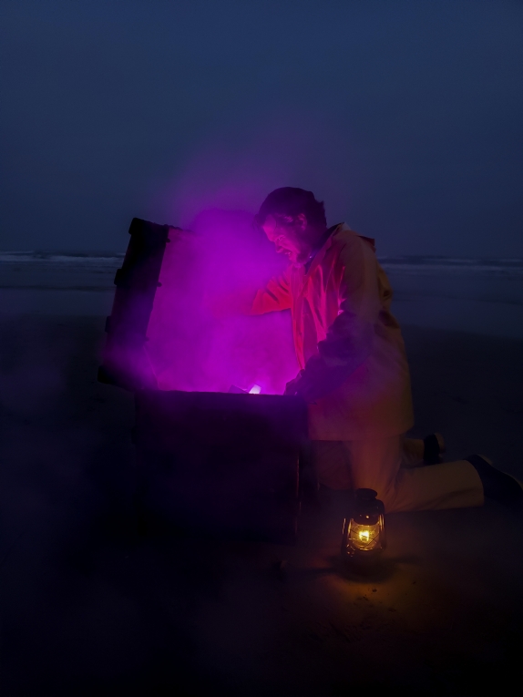 A bearded person in yellow rain gear kneels in profile on a dusky twilight beach lifting the lid of a chest that is emitting an intense violet light soaking into a rising fog. a glowing oil lamp rests at the person's knee.  