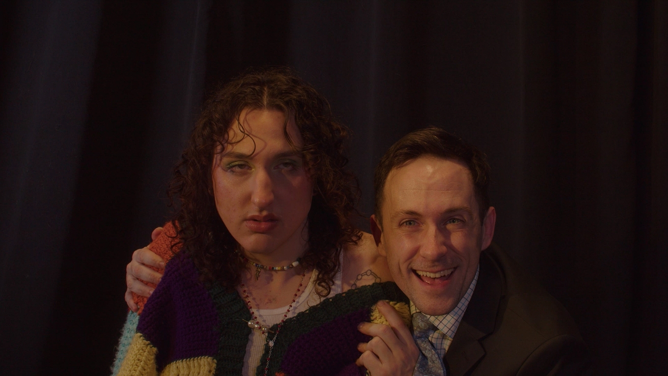 still from Be Thyself, with two people gazing directly past the viewer. the person on the left has long curle hair and is wearing a colorful knit cardigan. their face has an exhausted woozy look. a person in a suit and tie and a gleeful smile is coming up behind the first person with hands placed on each shoulder. 