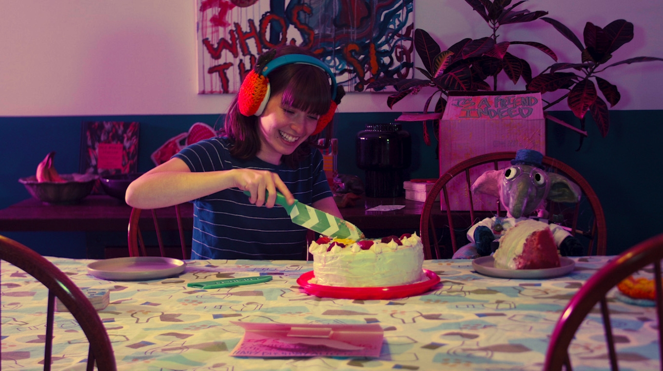 still from thank you for going goblin. a person in a striped blue tshirt and chunky red headphones gleefully slices into an iced cake using a green chevron-patterned knife at a quirky dining room tables next to an elephant plushie. the whole room is bathed in violet light. 