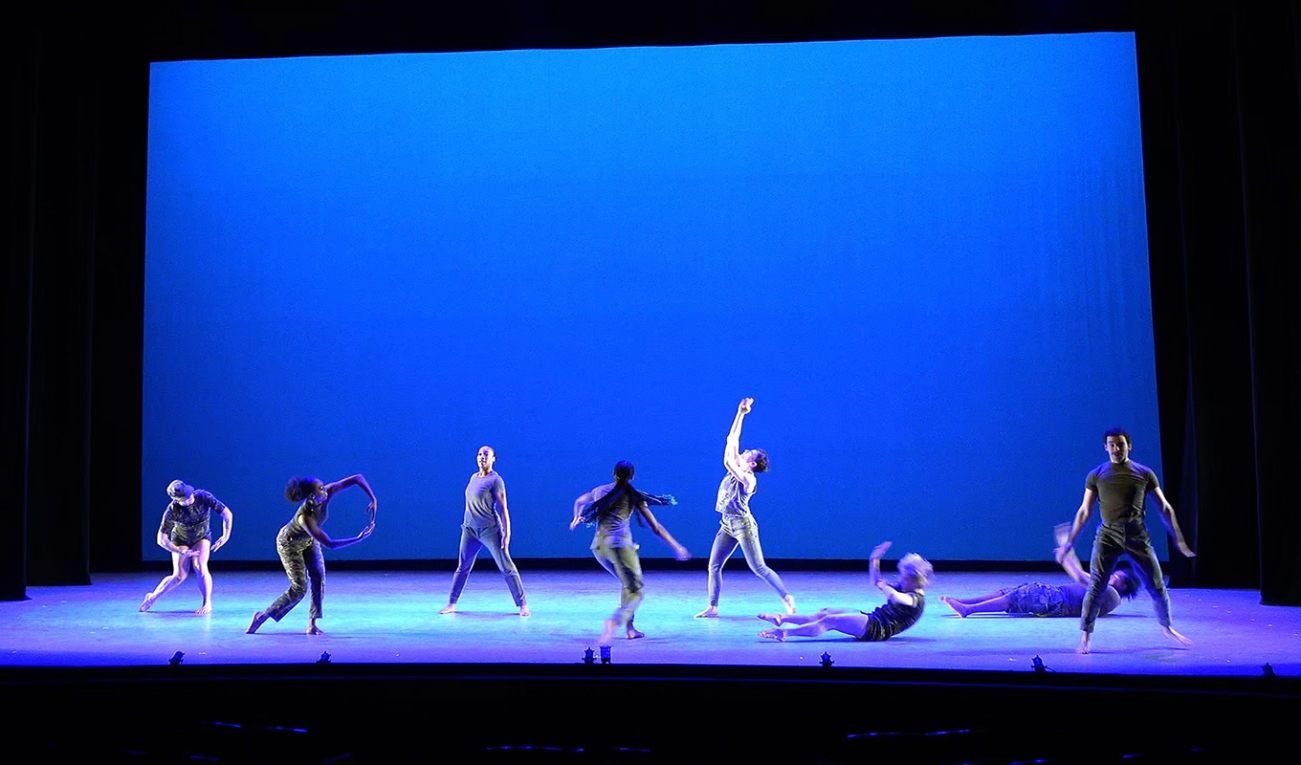 eight dancers are seen in various positions across a large stage illuminated in blue light. a blue rectangular screen illuminates the wall behind the dancers. some are making their arms into circles other are laying on the floor. 