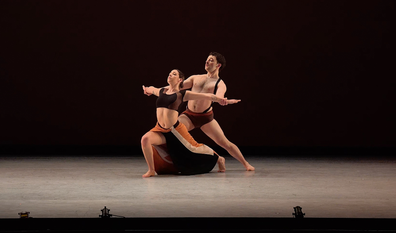 two dancers on a stage illuminated with a warm gray tone dance one in front of the other. the front dancer has an orange and black skirt and is lunging forward with arms held wide. the rear dancer has ochre briefs and is holding the other dancers arms. 