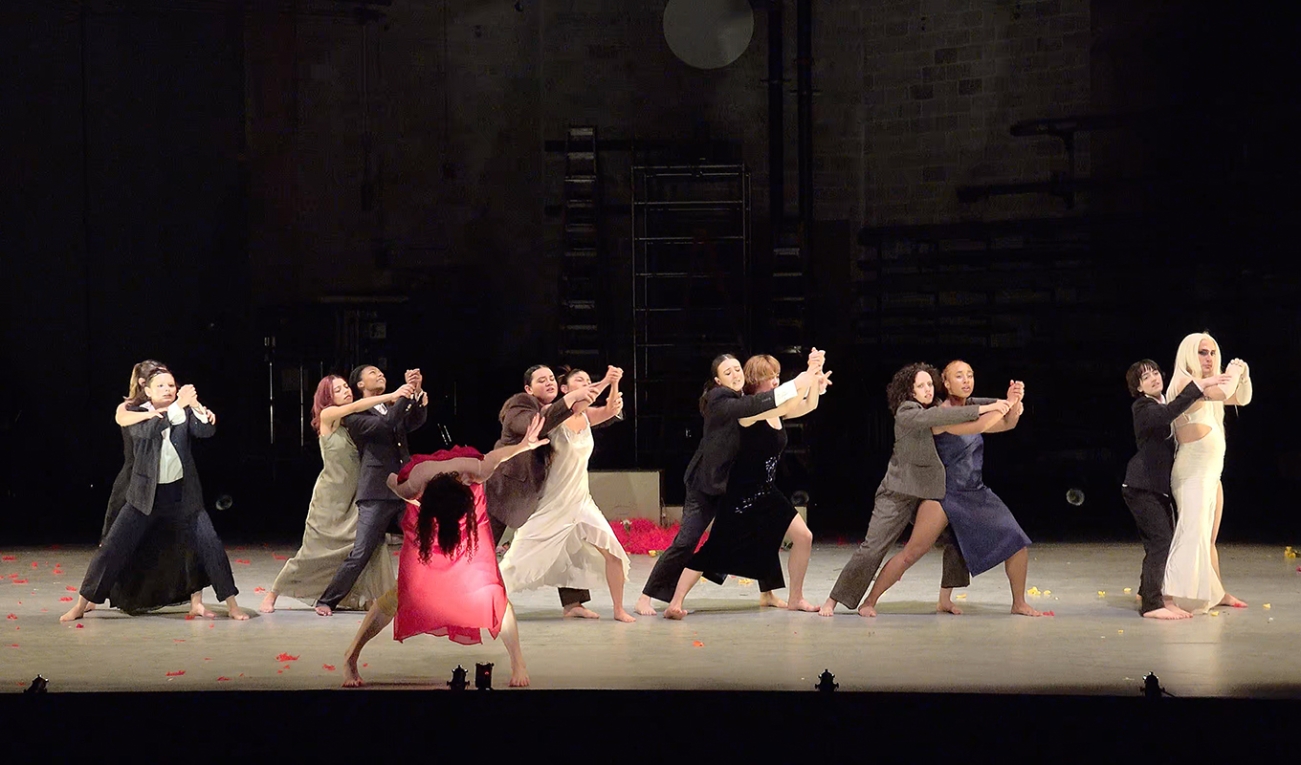 13 dancers are dancing in an industrial space with a brightly illuminated dancefloor. the six pairs consist of a dancer in a suit and a dancer in a dress. a thirteenth dancer in a red dress is bent over in the front left of the row of pairs. 