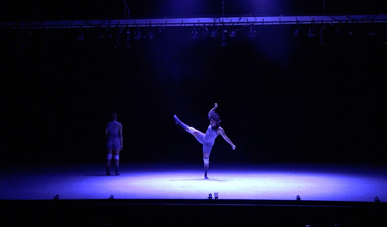 a dancer with a legged kicked high is in the center of a stage beneath a cold large bluish spotlight. another dancer stands with back facing the viewer at the edge of the spotlight.