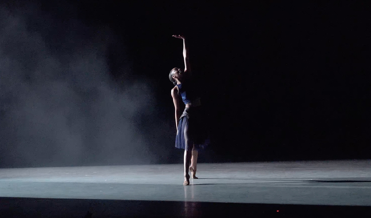 a single dancer in a black outfit with sheer black shirt dances in a fog-filled dancefloor illuminated from the side with white light, one hand stretched up high into the beam of light. 