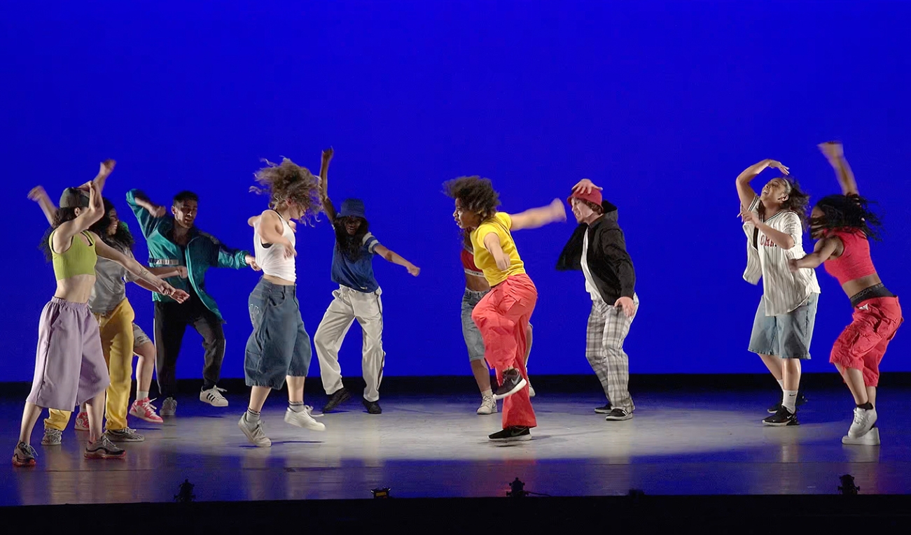 ten dancers in casual, vaguely 90s style attire perform under a white spotlight on a stage, with a rear wall illuminated in a deep blue. 