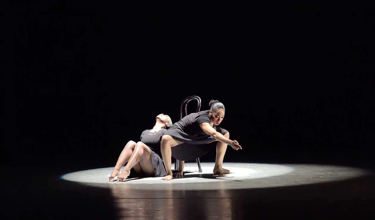 two dancers perform in a narrow, bright spotlight on an otherwise completely black stage. one person is seated gazing upwards, the other crouching in a deep stance with an arm outstretched. 
