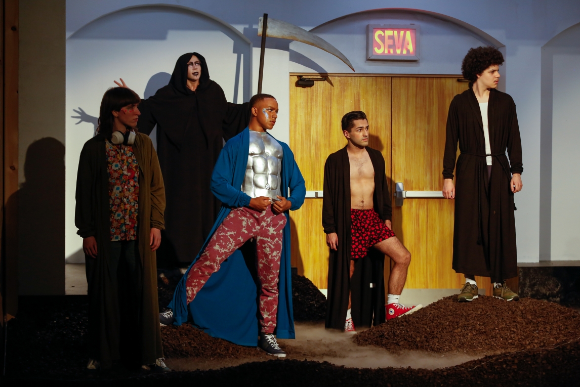 five actors stand in a spotlight beam on a stage of rolling mounds of dirt, all gazing to the photo's right. from right to left, their costumes include a brown bathrobe, a wide open brown bathrobe and red boxer shorts, bottle pink pants with a silver armor breastplate, a hooded grim reaper outfit with a scythe, an olive bathrobe and floral button-up. 