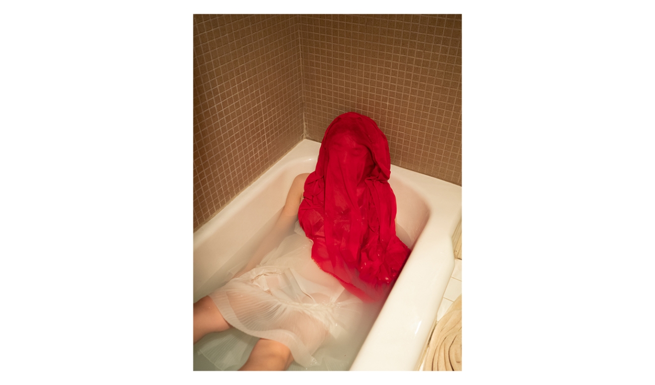 photographic work depicting a person ins a sheer white dress in a filled bathtub with a crimson sheet over their face and torso. flat brownish square tiling lines the walls around the person slumping back. 