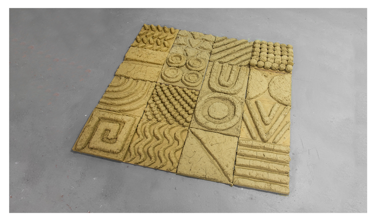 a sculptural artwork of sixteen yellowish tiles each with a different texture and design. 