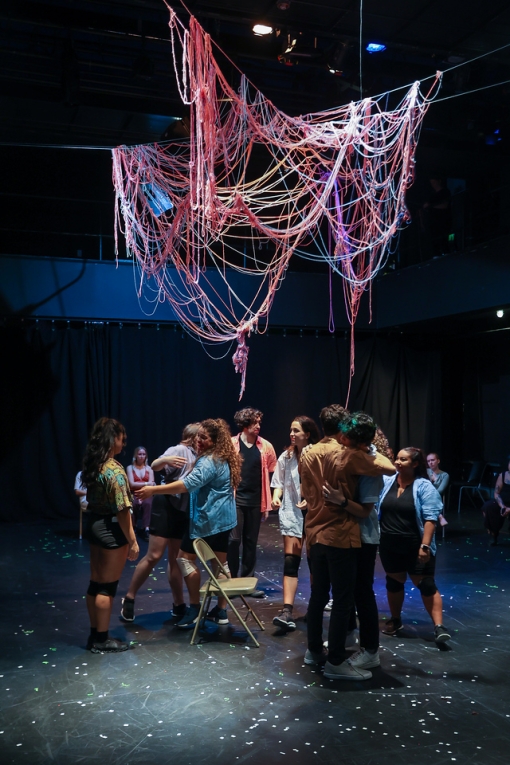 Inside of a black box style theater, a massive tangle of reddish yarns is suspended above a cast of actors milling around and embracing. the tangle of yarn dips low to the ground and is looping up and down.  