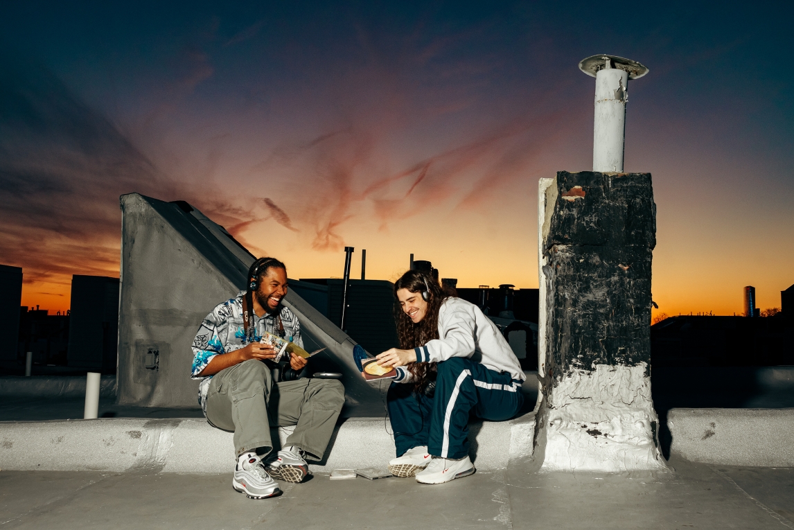 two people sitting on a rooftop ledge smiling and laughing while wearing headphones and examining CDs against a sunset sky that's quickly fading to dusk. 