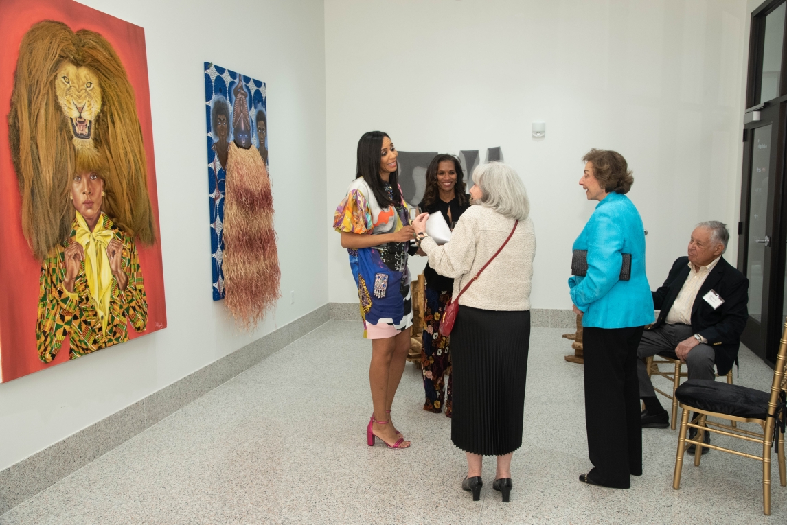 a group of people converse in a gallery space, with large paintings on the wall at the left and people huddled up in the center. at the right are two chairs painted gold, with one person seated on the further one from the viewer 